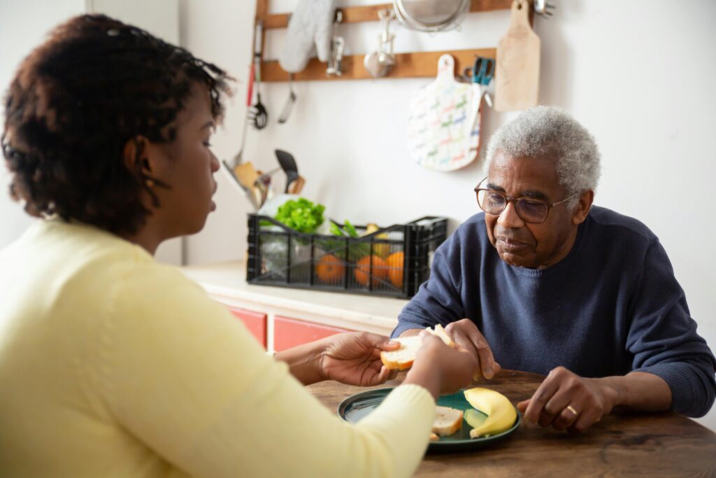 A woman assists an elderly man with meals at home, showcasing care and support.